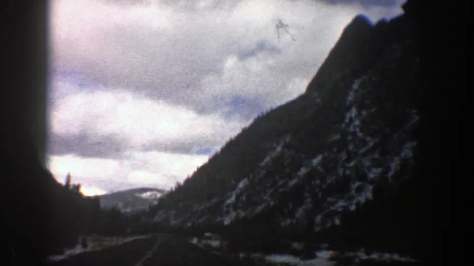 1961 Aspen-area valley view showing dark mountain slopes, a road-like corridor below, distant snow, and gray-white clouds.