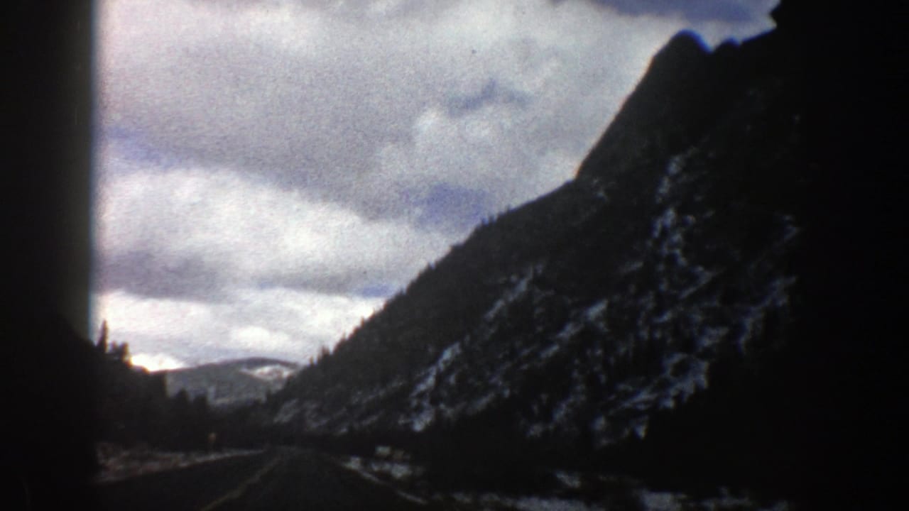 Mountain road beneath cloudy sky with a dark snow-dusted slope on the right.