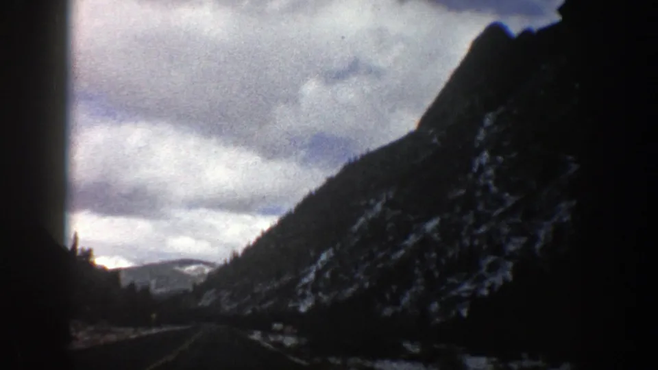 1961 Aspen, Colorado 8mm frame showing a forward road view beside a steep snow-dusted mountain slope under clouds.
