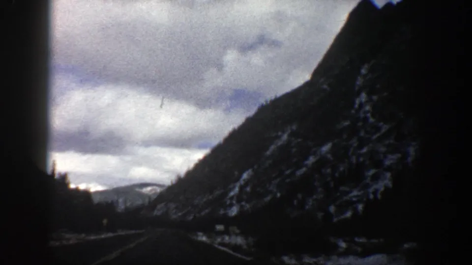 Forward-facing 1961 Aspen travel frame with a roadway, evergreen-covered slope, snow patches, and overcast mountain sky.