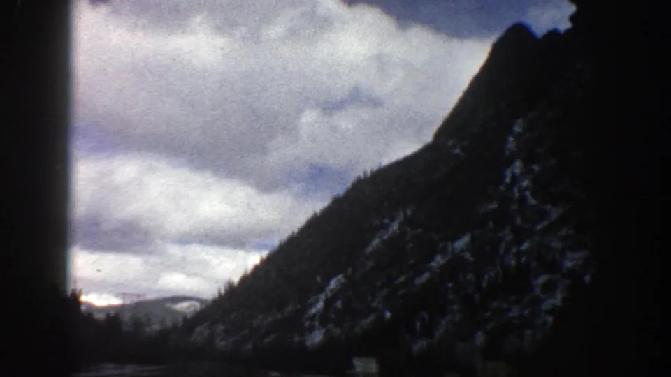 Late frame from the 1961 Aspen 8mm clip showing a road through mountains, a steep snowy slope, and layered clouds.