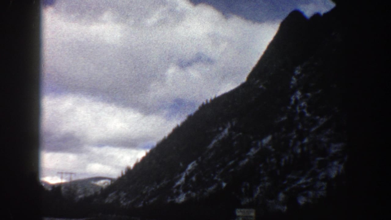 Mountain road view through a car windshield with dark rock slope, clouds, and snowy ridges.