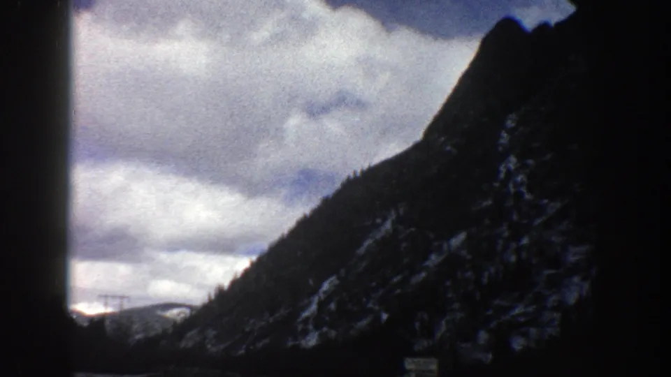1961 Aspen, Colorado home-movie frame looking through a windshield toward a mountain road, rocky slope, evergreen trees, clouds, and distant snow.