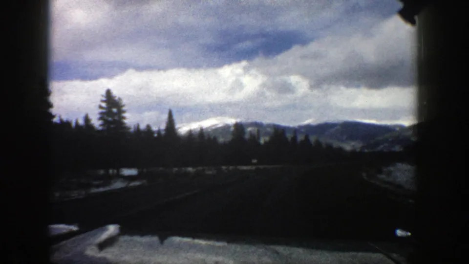 1961 Aspen, Colorado windshield view of a curving road bordered by evergreens and snow patches, with distant mountains beneath layered clouds.