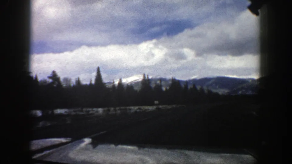 1961 Aspen, Colorado travel frame showing a windshield view across roadside snow, dark trees, distant mountains, and a bright cloudy sky.