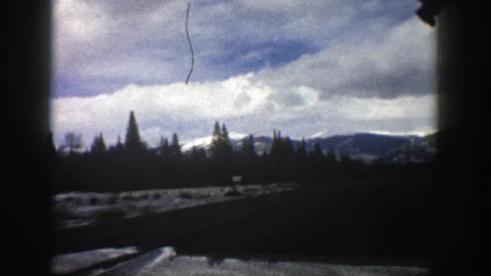 1961 Aspen, Colorado windshield shot with a dark roadway, roadside snow, evergreen silhouettes, distant mountain ridges, and cloudy sky.