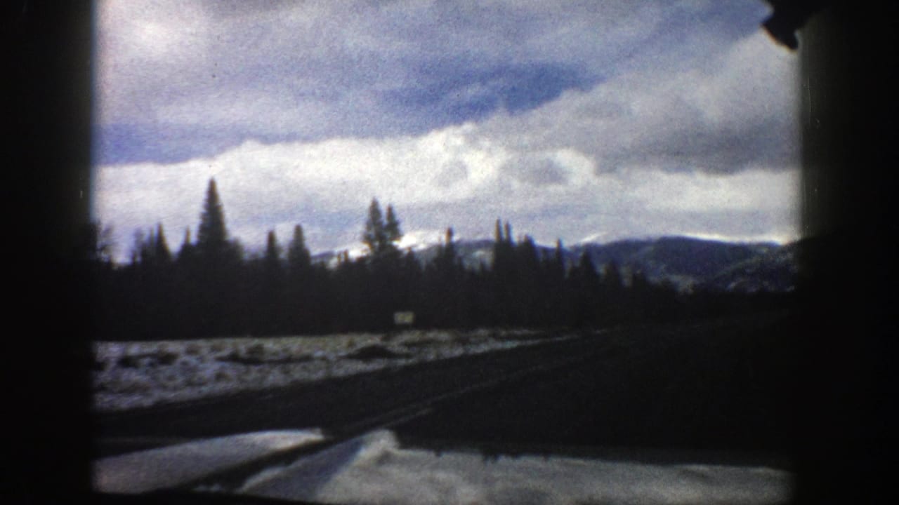 Vehicle-window view of a snowy mountain road with evergreen trees and distant ridges under cloudy sky.