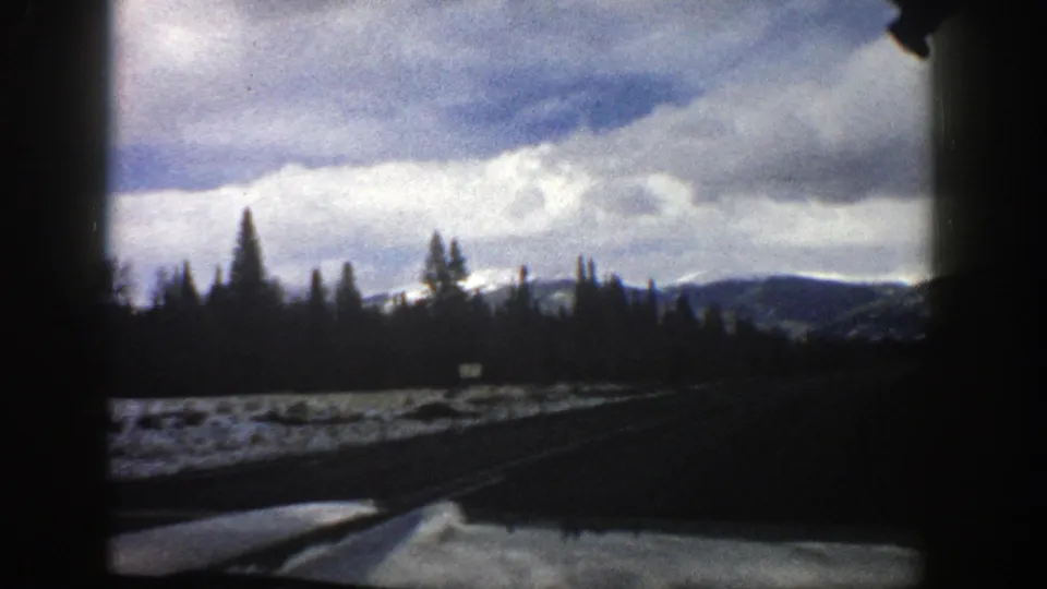 1961 Aspen, Colorado home-movie frame from inside a vehicle, showing a curving snowy road, dark dashboard edge, evergreens, and distant mountains.