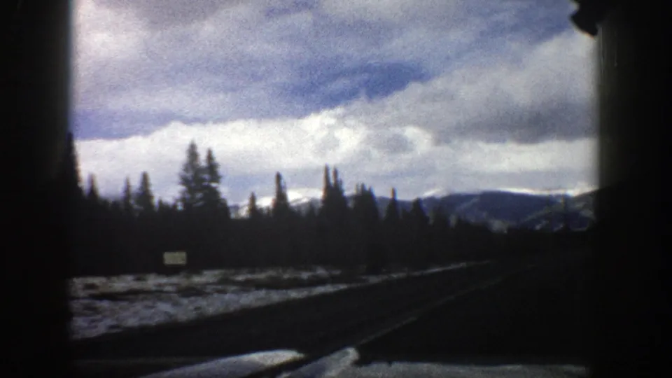 1961 Aspen, Colorado driving view with the road angling past snow patches, evergreen silhouettes, a small roadside sign, and low mountains beneath clouds.