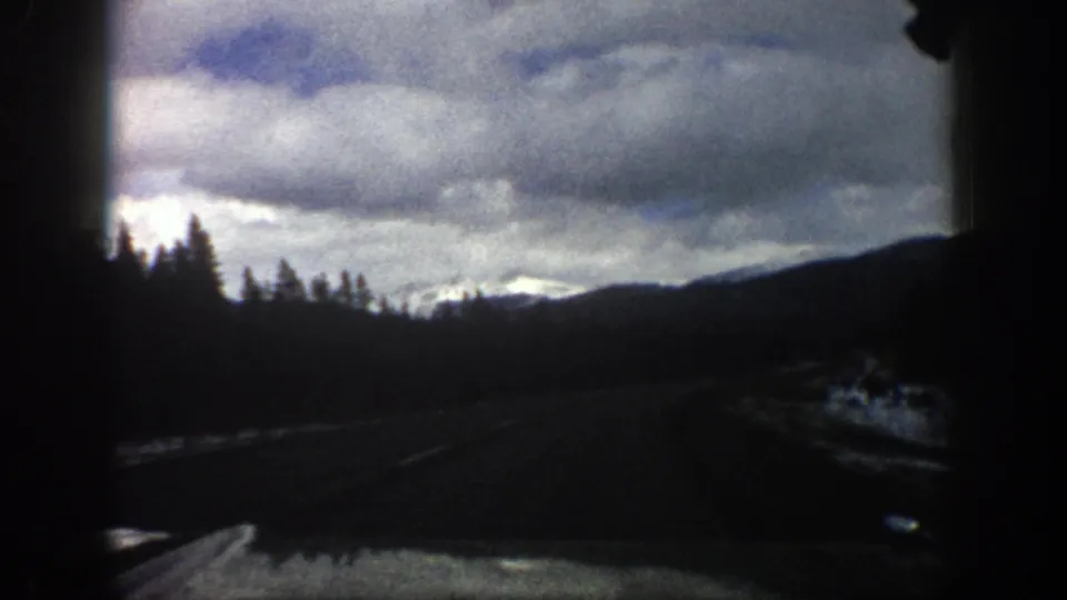 1961 Aspen, Colorado home-movie frame showing a dark in-car foreground, roadside trees, a mountain ridge, snow on the ground, and a dramatic cloudy sky.