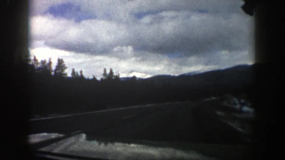 1961 Aspen, Colorado in-car travel frame with a curving road, snowy shoulder, evergreen tree line, distant mountain slope, and dense cloud cover.