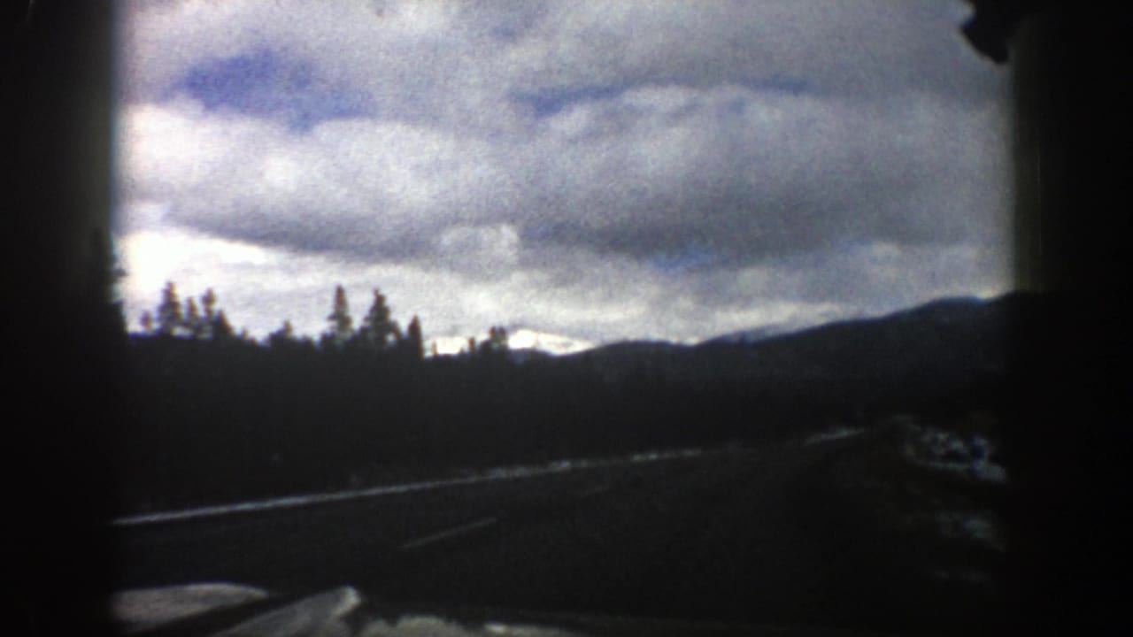 1961 Aspen, Colorado 8mm road-trip view from a moving car, showing a road edge, evergreens, distant ridges, and heavy clouds.