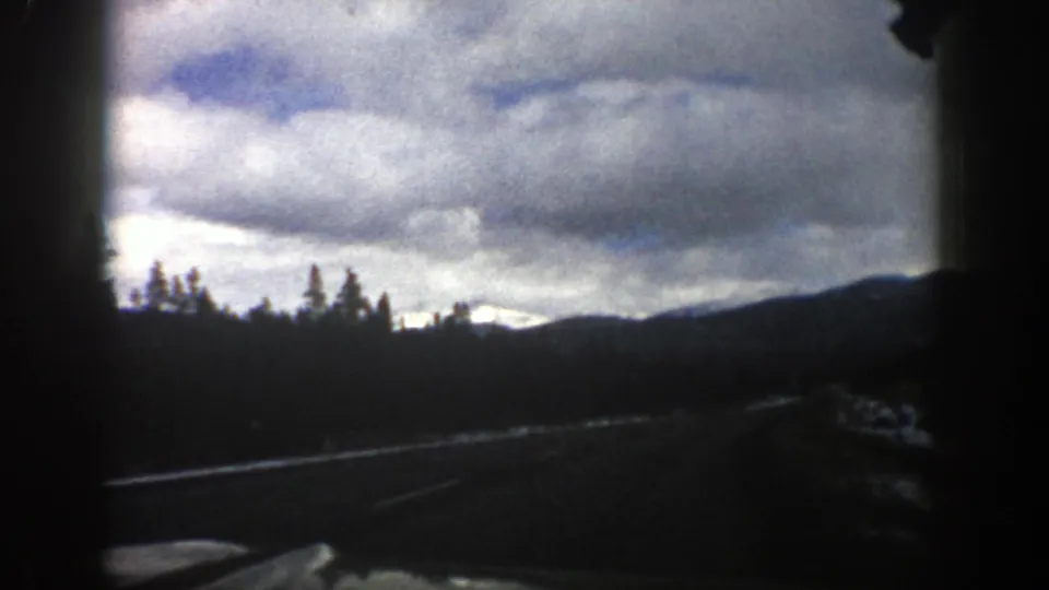 1961 Aspen, Colorado 8mm road-trip view from a moving car, showing a road edge, evergreens, distant ridges, and heavy clouds.