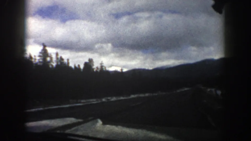 1961 Aspen, Colorado home-movie frame showing a roadside view of evergreens, snow patches, dark mountain ridges, and dramatic clouds.