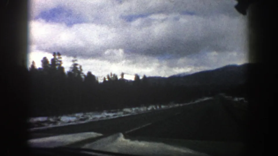 1961 Aspen, Colorado 8mm frame of a curving mountain road, roadside snow, evergreen trees, and overcast sky from a moving car.