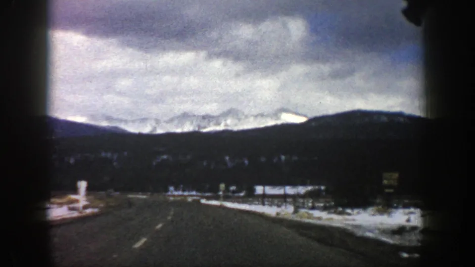 1961 Aspen, Colorado road-trip frame with a paved road leading toward snowy peaks, dark hills, roadside snow, and a cloud-filled sky.