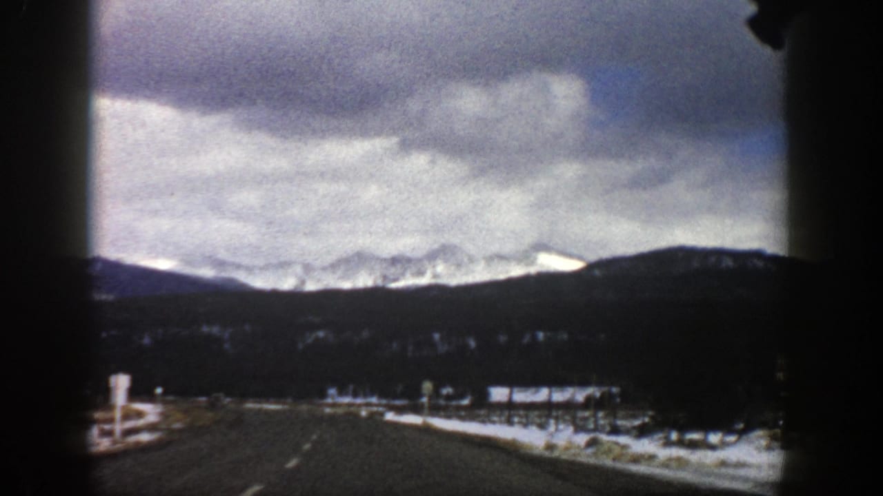 1961 Aspen-area 8mm view from a moving car on a winter highway, with roadside snow, dark forested hills, and distant mountains.