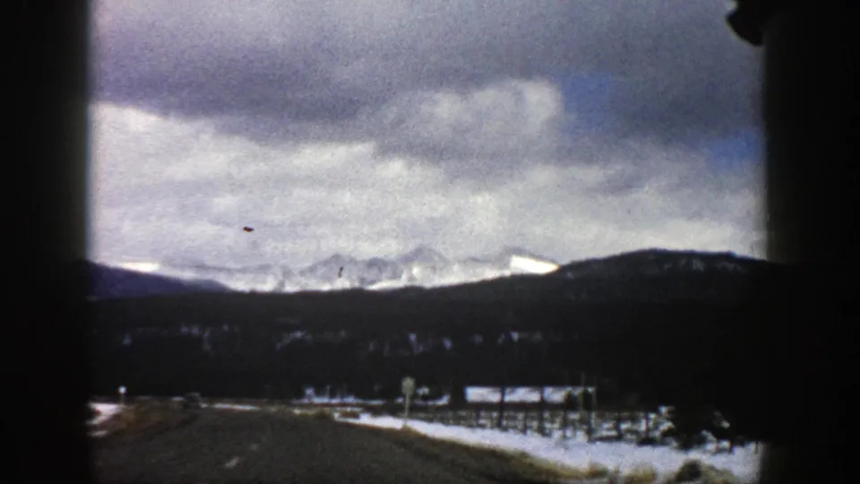 1961 Colorado winter road view with snow patches beside the highway, dark evergreen slopes, and pale mountains in the distance.