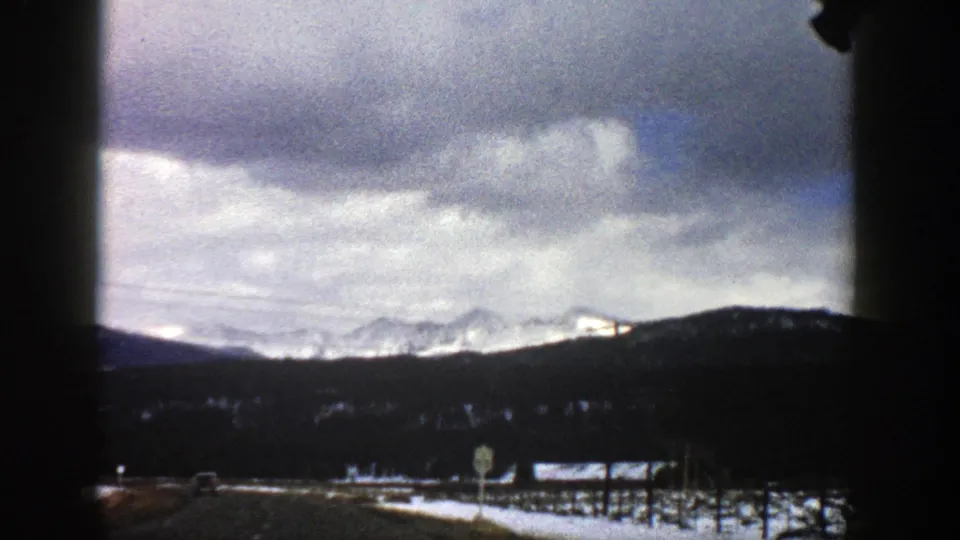 Aspen-area winter highway footage from 1961, looking ahead past road signs and snow toward dark hills and snow-capped peaks.