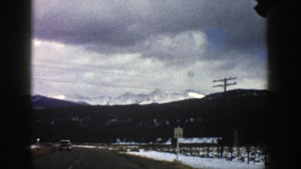 1961 Aspen, Colorado road scene with a distant car, utility poles, snowy roadside, dark hills, and mountains under overcast clouds.