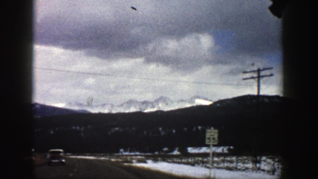 1961 Colorado 8mm frame from a moving car showing a roadside speed sign, utility poles, snow patches, and distant mountains.