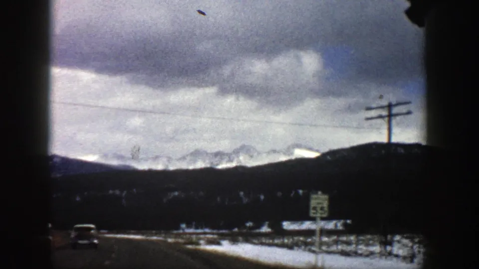 1961 Colorado 8mm frame from a moving car showing a roadside speed sign, utility poles, snow patches, and distant mountains.