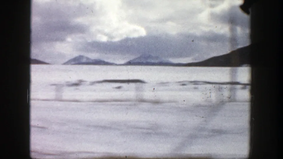 1961 Colorado travel frame showing a wide frozen or pale lake, low shoreline shapes, mountains, and car-window framing.