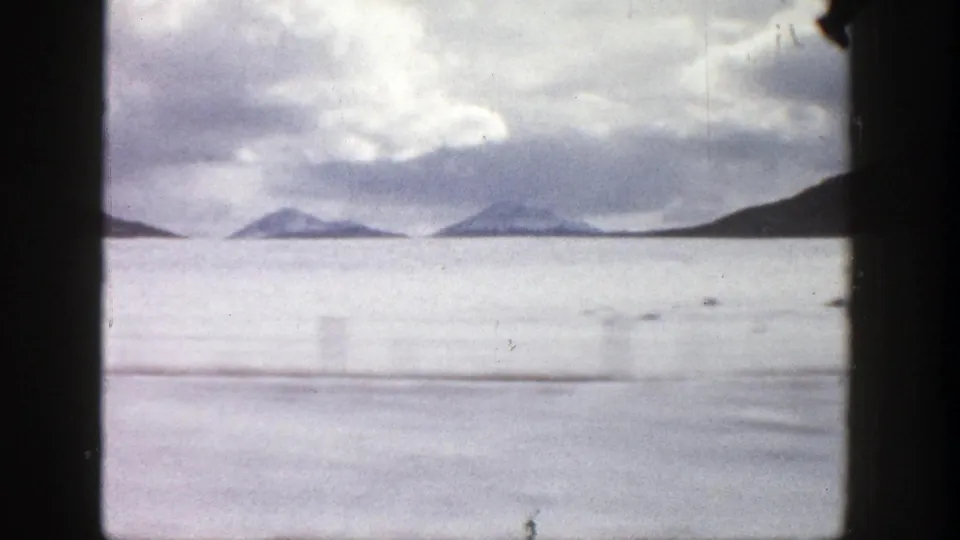 1961 Colorado 8mm frame with a frozen lake or pale water, distant mountain ridges, and heavy overcast sky seen from a car.
