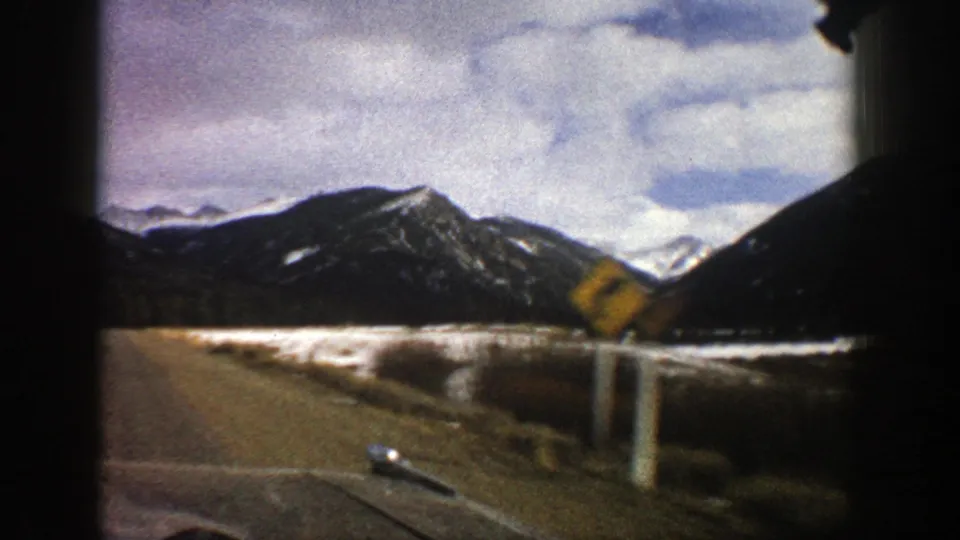 Moving-car view near Aspen in 1961 showing a road edge, snow-dusted mountains, roadside posts, and a yellow warning sign.