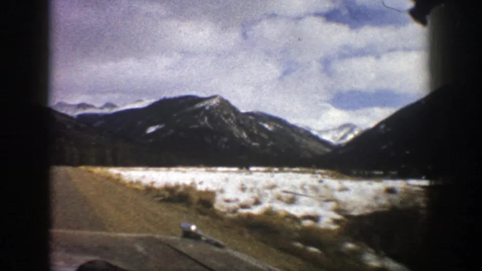 1961 Aspen-area road-trip frame showing a curving road, snowy roadside, dark mountain ridge, and soft 8mm color.