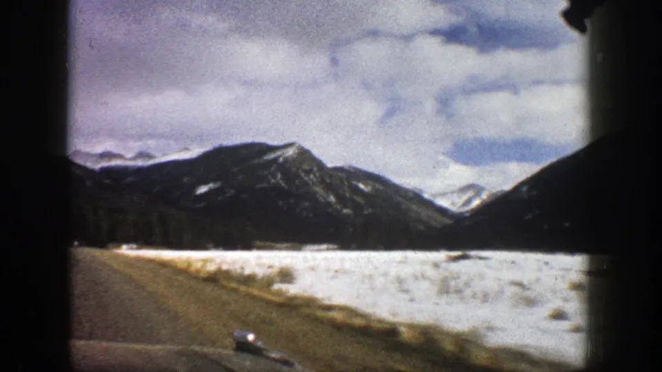 8mm home-movie frame from 1961 Aspen showing a snowy roadside valley and mountain backdrop from a passenger viewpoint.