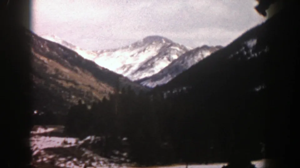 1961 Aspen, Colorado 8mm view of snow-covered mountain peaks rising over a dark wooded valley under a pale sky.
