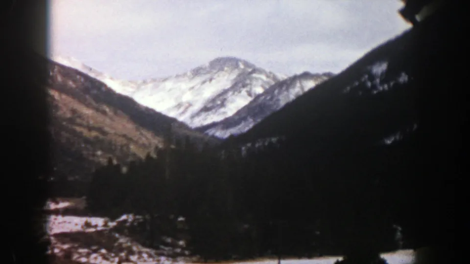 1961 Aspen-area 8mm frame showing snowy peaks, evergreen forest, and dark gate-edge shadows from a moving-view mountain shot.
