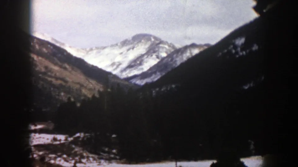 1961 Aspen, Colorado home-movie frame with distant snow-covered ridges, evergreen trees, cloudy sky, and visible 8mm edge shadow.