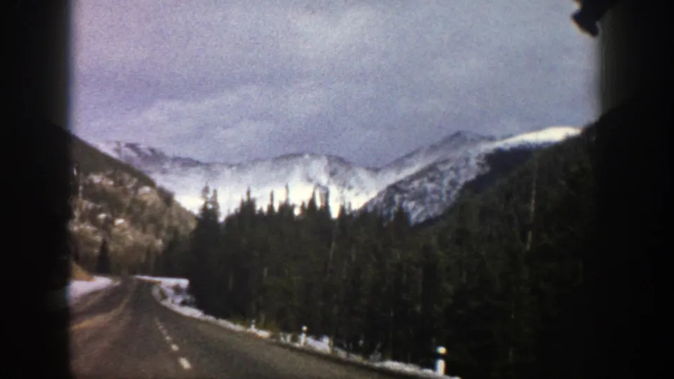 1961 Aspen-area travel frame showing a paved mountain road through evergreens toward snow-covered peaks under a cloudy sky.