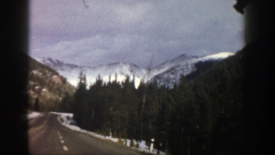 1961 Aspen, Colorado 8mm frame of a winding road, roadside snow, dense evergreens, and distant snowy mountains.
