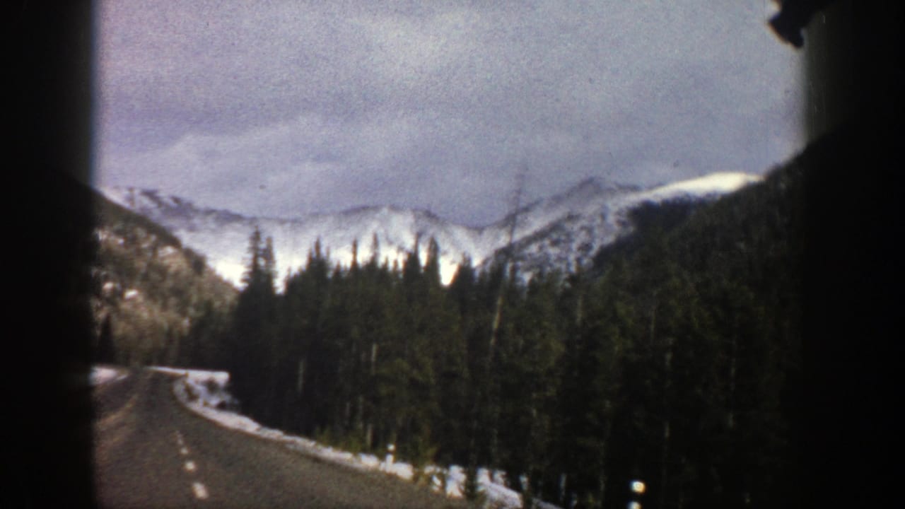 Two-lane mountain road curving past evergreens toward snowy peaks under a cloudy sky.