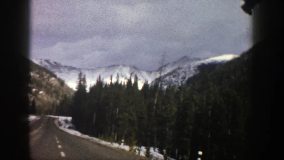 1961 Aspen, Colorado 8mm view of a road curving through evergreen forest toward snow-covered mountains.