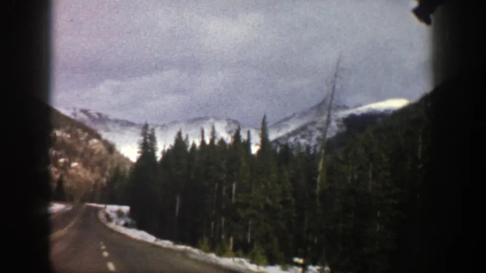 Aspen-area road view from a moving vehicle, with snowbanks, pine forest, and distant mountains in 1961 8mm footage.