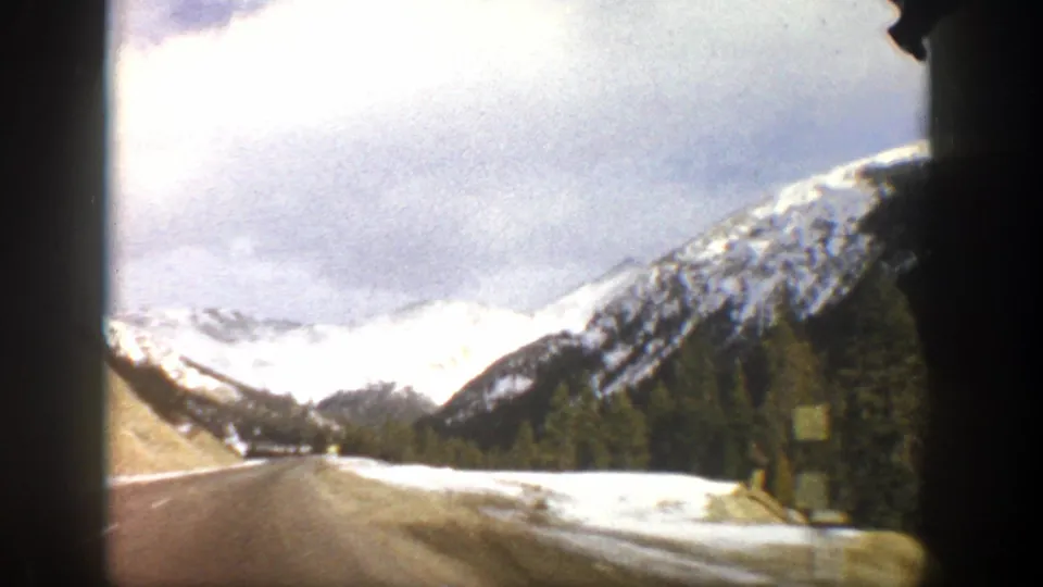 1961 Colorado mountain-drive frame showing a paved road, roadside snow, green trees, and bright snow-covered peaks.
