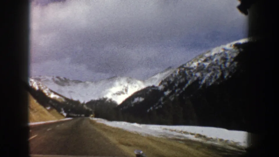 Aspen, Colorado 8mm travel view with a road framed by snowbanks, evergreen forest, and cloud-covered mountain peaks.