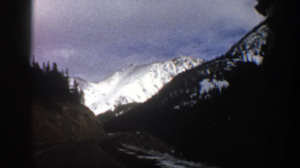 1961 Aspen, Colorado travel frame with a bright snowy peak framed by dark evergreen hills and cloudy sky.