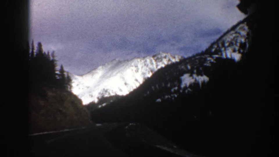 1961 Aspen, Colorado 8mm view from a vehicle on a dark mountain road below a snow-covered peak.