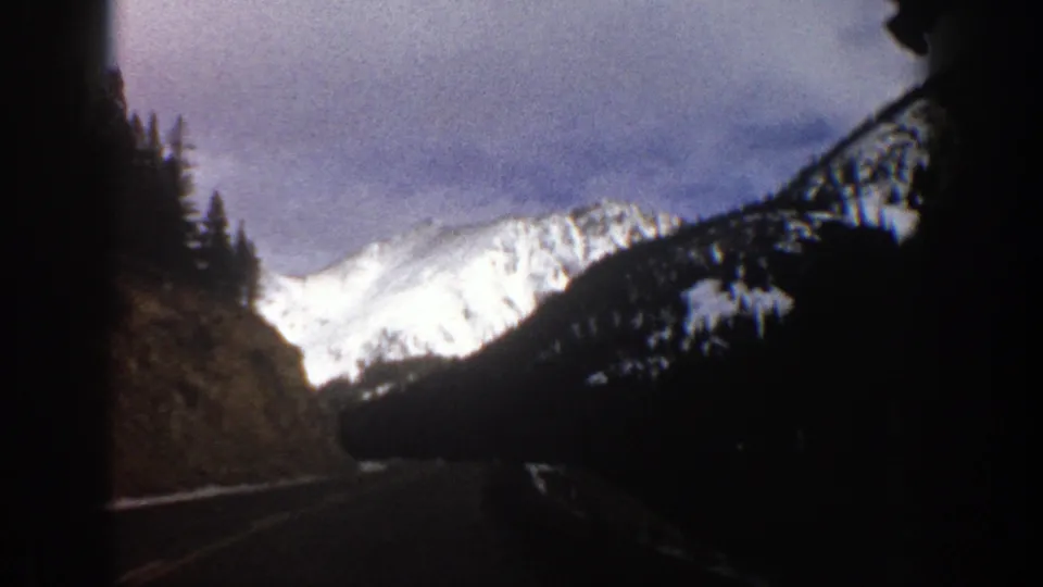1961 Aspen, Colorado frame showing a snowy mountain beyond a shadowed road and evergreen slopes.