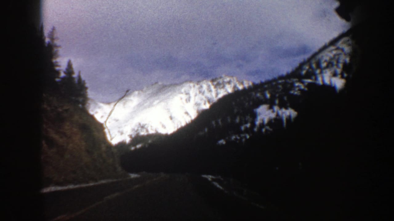 1961 Aspen-area 8mm frame from a moving vehicle, showing a dark mountain road leading toward snowy peaks and forested slopes.