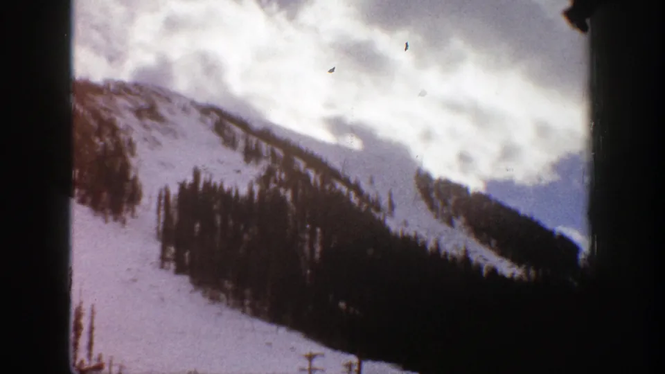1961 Aspen, Colorado home-movie frame with a snow-covered ridge, dense evergreens, cloudy sky, and visible 8mm frame borders.