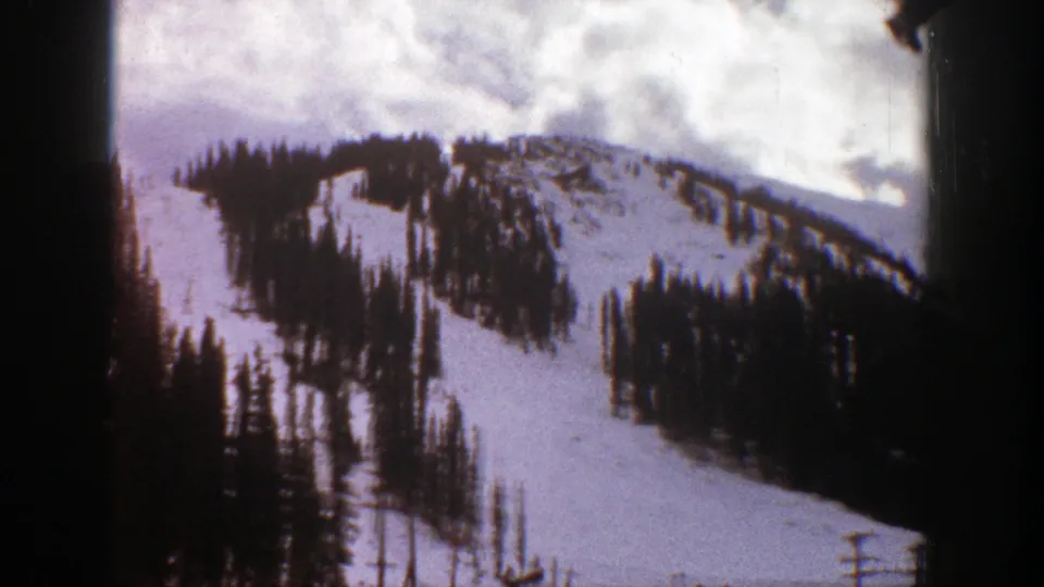 1961 Aspen, Colorado frame showing a snow-covered mountainside, dark evergreen stands, and bright clouds within the 8mm film frame.