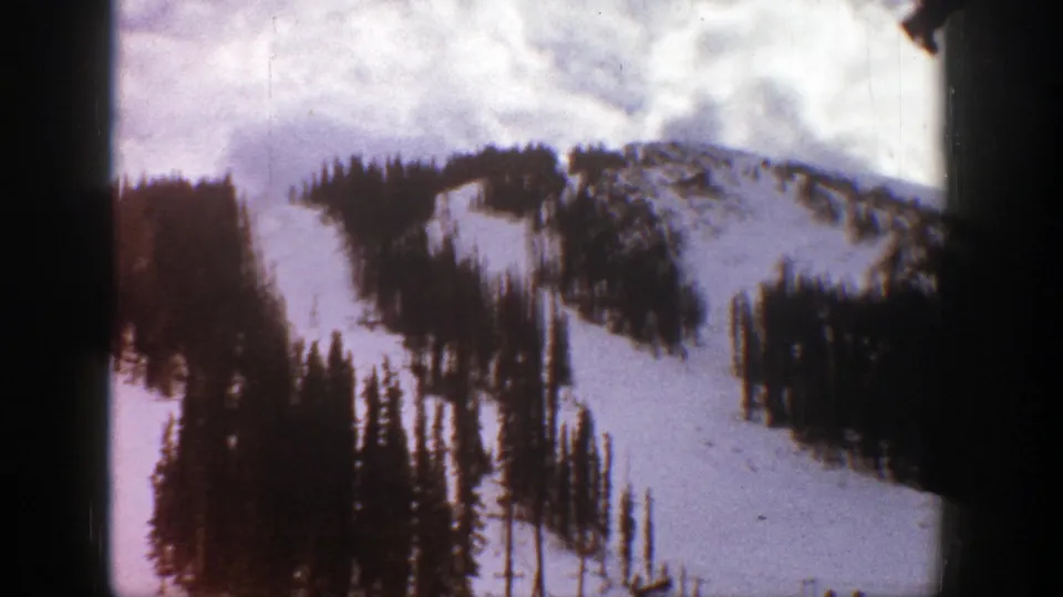 1961 Aspen home-movie frame showing snowy ski runs winding through evergreen forest beneath a cloudy mountain sky.