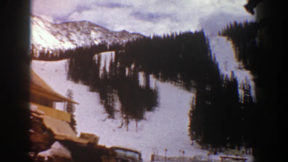 1961 Aspen home-movie frame showing snowy slopes, dense evergreens, distant mountains, and a small roofline at the bottom.
