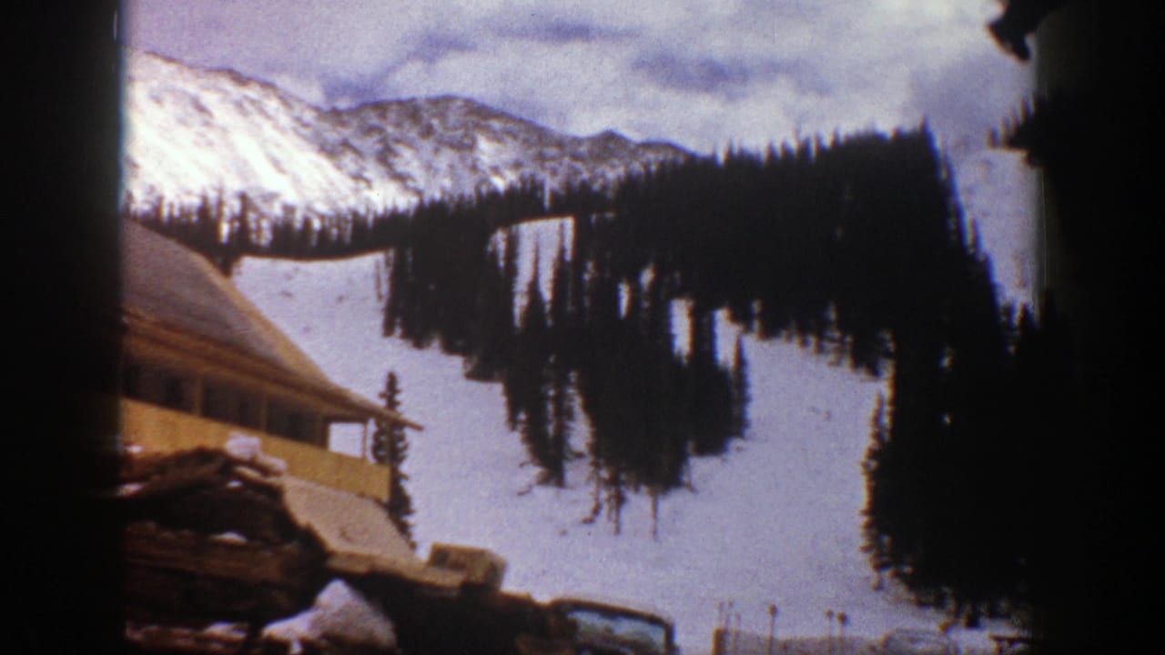 1961 Aspen home-movie frame showing a snow-covered slope, dark evergreens, distant ridge, and the edge of a rustic building.